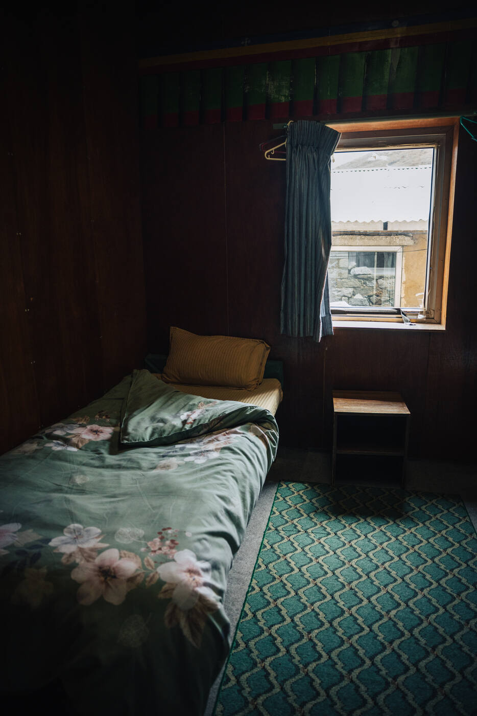 A simple lodge bedroom with green floral bedding, wood-paneled walls, and a small curtained window