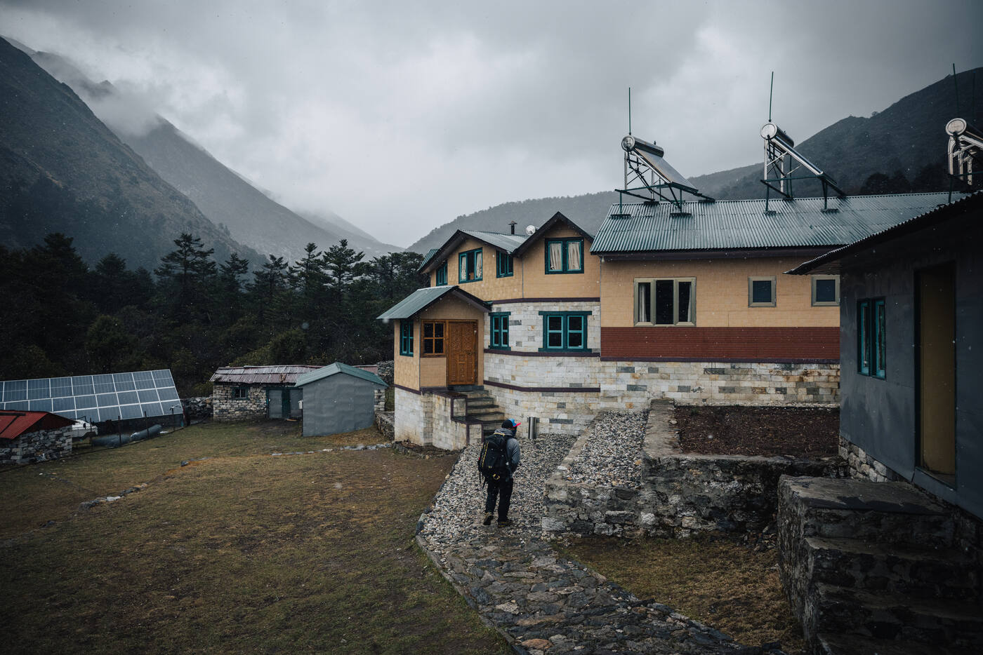A trekker walking up to a yellow stone lodge with solar water-heating panels on the roof, pine trees and foggy peaks behind