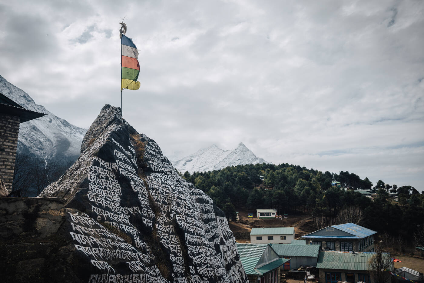 A carved mani rock topped by a striped Buddhist flag on the ridge above Namche, snow peaks behind