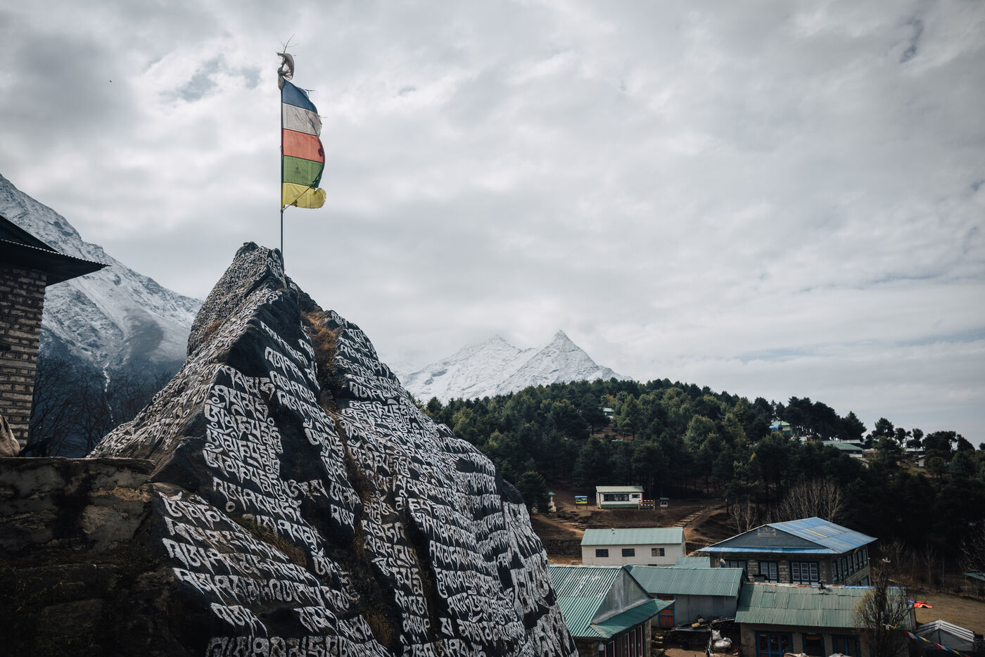A massive mani rock carved with white Tibetan script and topped with a single prayer flag, lodges and snow peaks behind