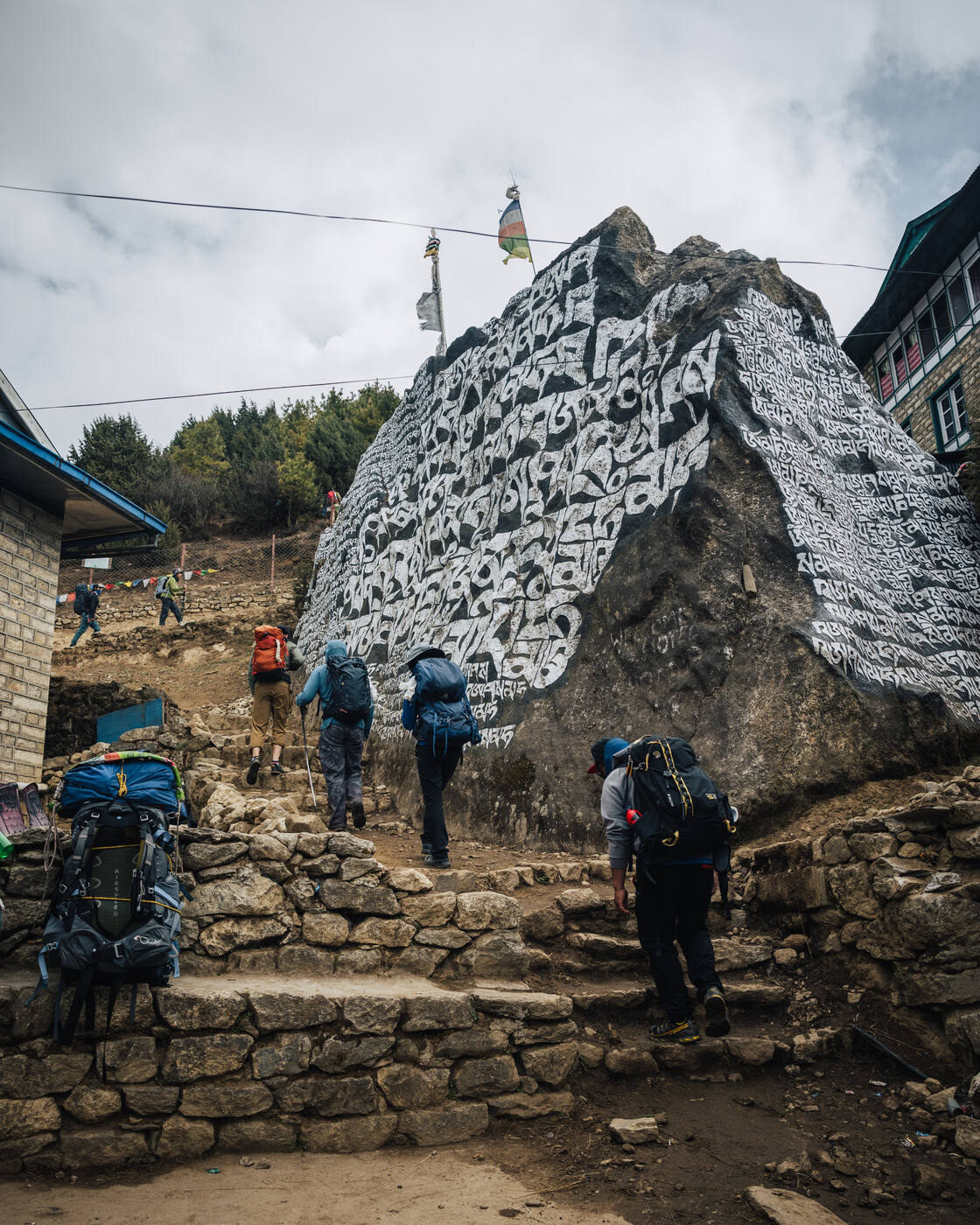 Trekkers ascending stone steps past a massive painted mani boulder, prayer flags strung above
