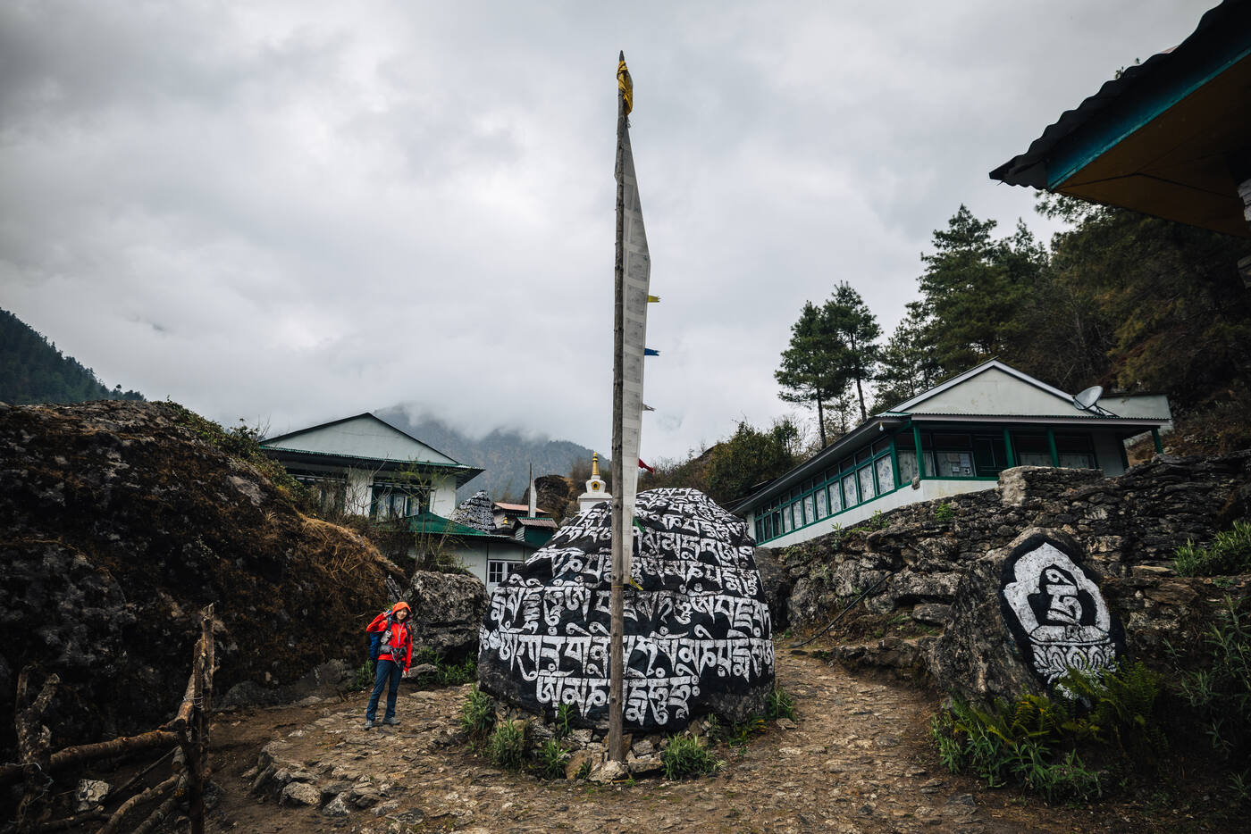 A trekker in red beside massive painted mani stones with Tibetan mantras, tin-roof lodges behind under a misty sky