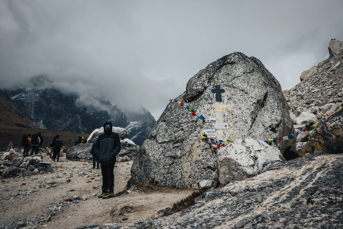 Trekker beside a massive memorial boulder etched with a Christian cross and trimmed with prayer flags — a private memorial on the Thukla ridge