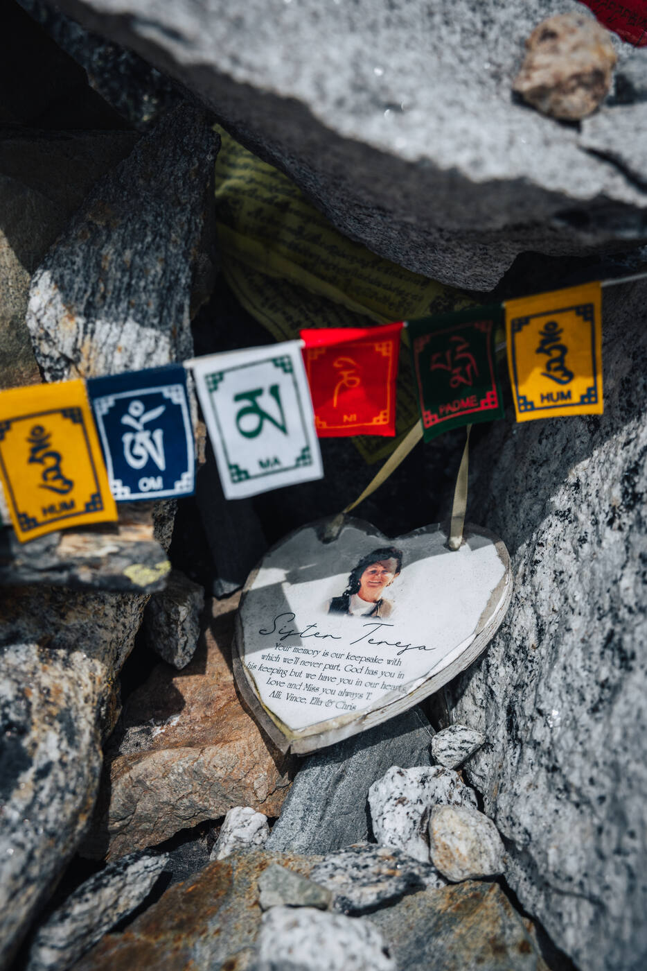 Close-up of a heart-shaped memorial plaque tucked among stones with a smiling photo and prayer flags above — a personal Chukpi Lhara tribute