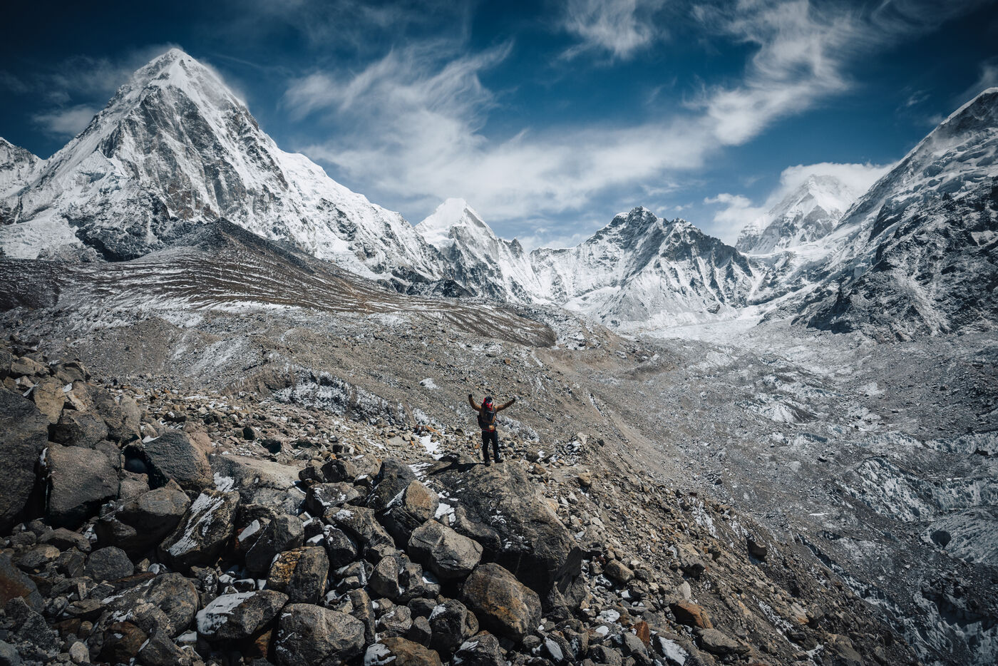 Trekker on a jumbled moraine with arms raised, Pumori's sharp pyramid on the left and a panorama of snow peaks spreading to the horizon