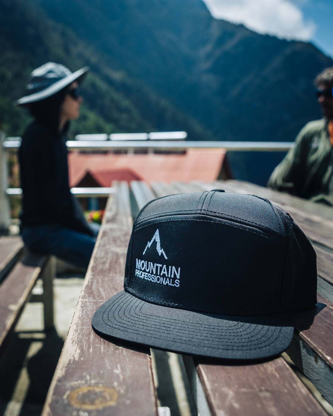 A black Mountain Professionals ball cap resting on a wooden bench with two trekkers and a peak in the background