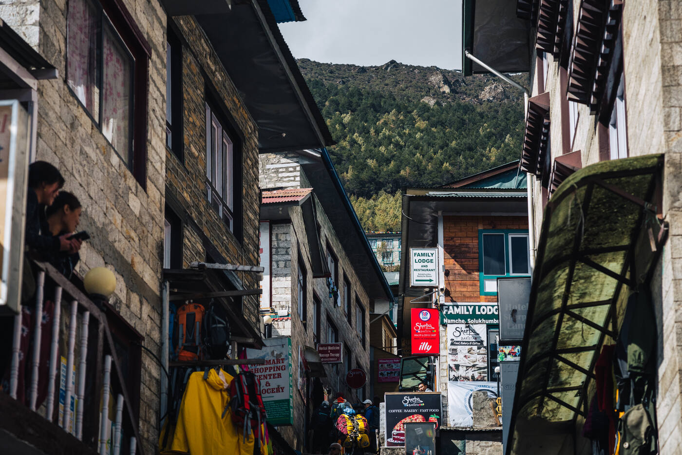 A narrow Namche alley lined with gear shops — Family Lodge sign, yellow rain jackets hanging outside