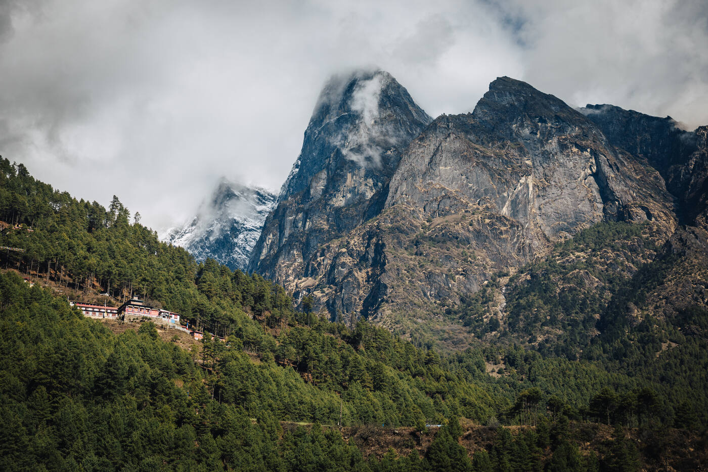 A dramatic rocky peak with cloud-wrapped summit rising above forested slopes, a small lodge perched on the ridge