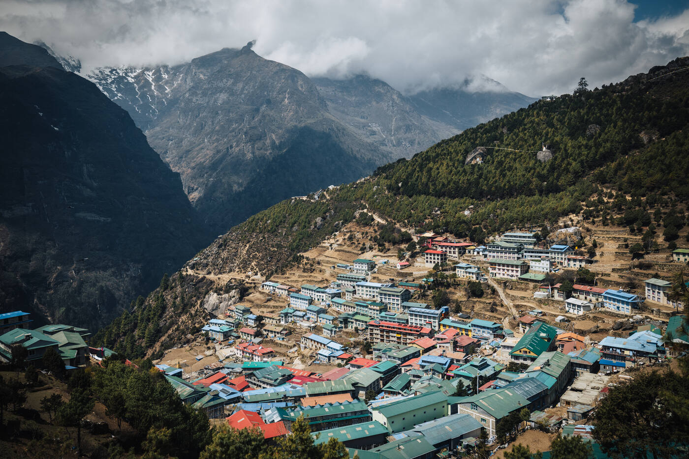 Namche Bazaar seen from above, clustered green and blue rooftops with peaks rising behind