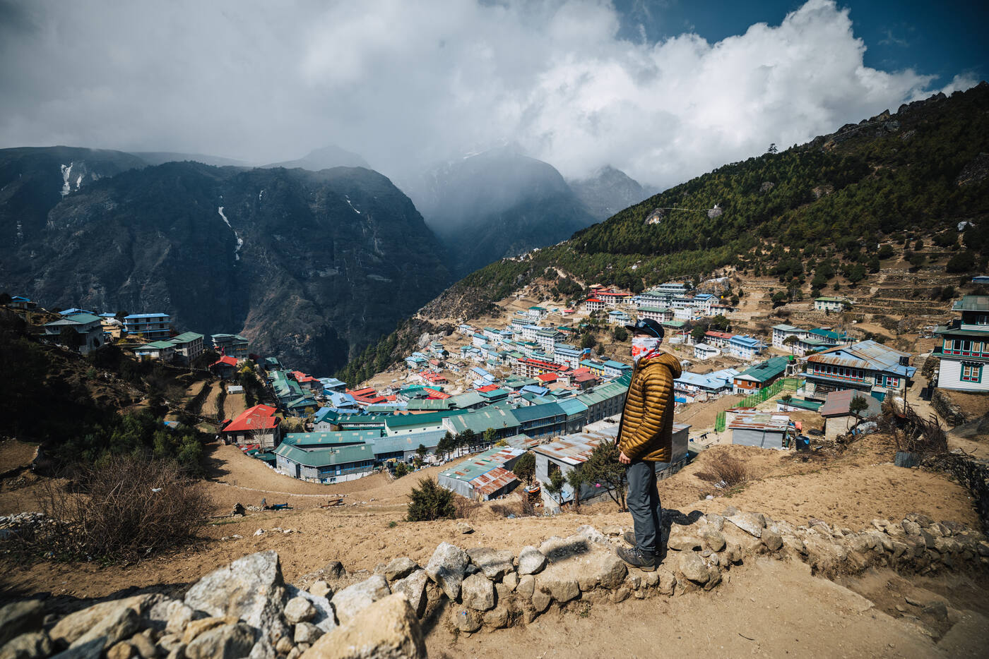 A trekker in a yellow puffer looking out over the Namche bowl from a rocky viewpoint above town