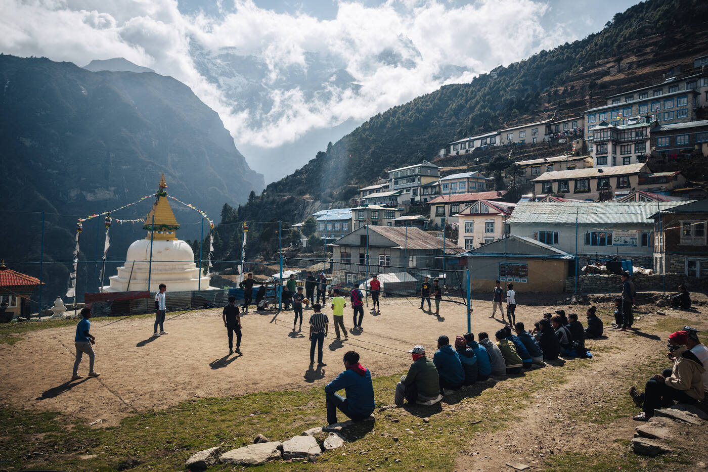The Namche main square — villagers gathered around a sports court with the tall white stupa and Namche bowl rising behind