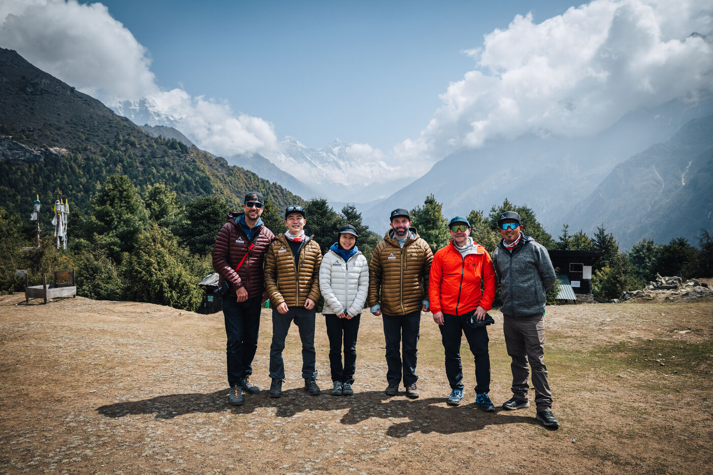 Six trekkers standing together with Himalayan peaks and prayer flags behind at the viewpoint above Namche