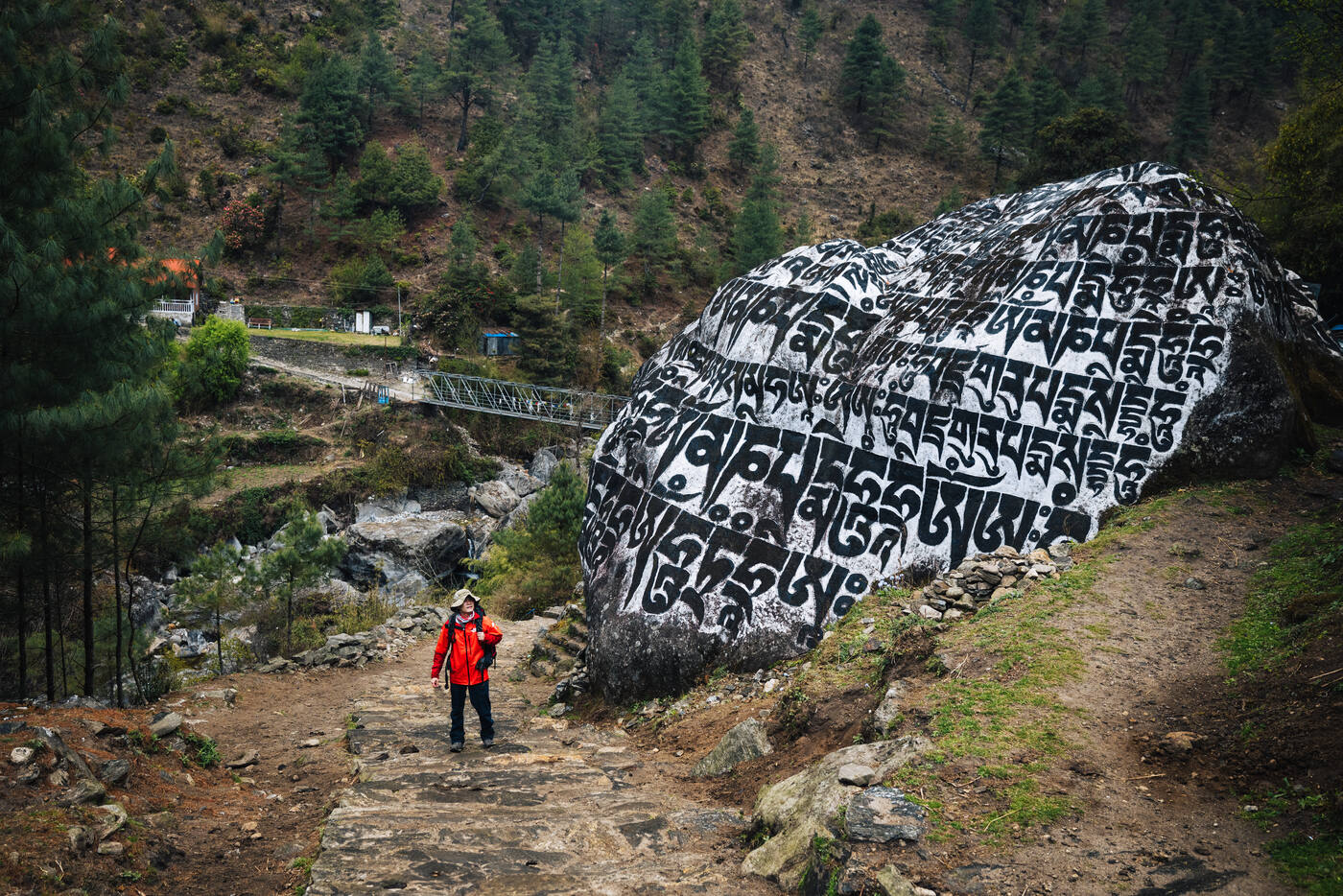 Trekker in a red jacket walking past a massive painted mani boulder covered in white Tibetan mantras, a small bridge behind