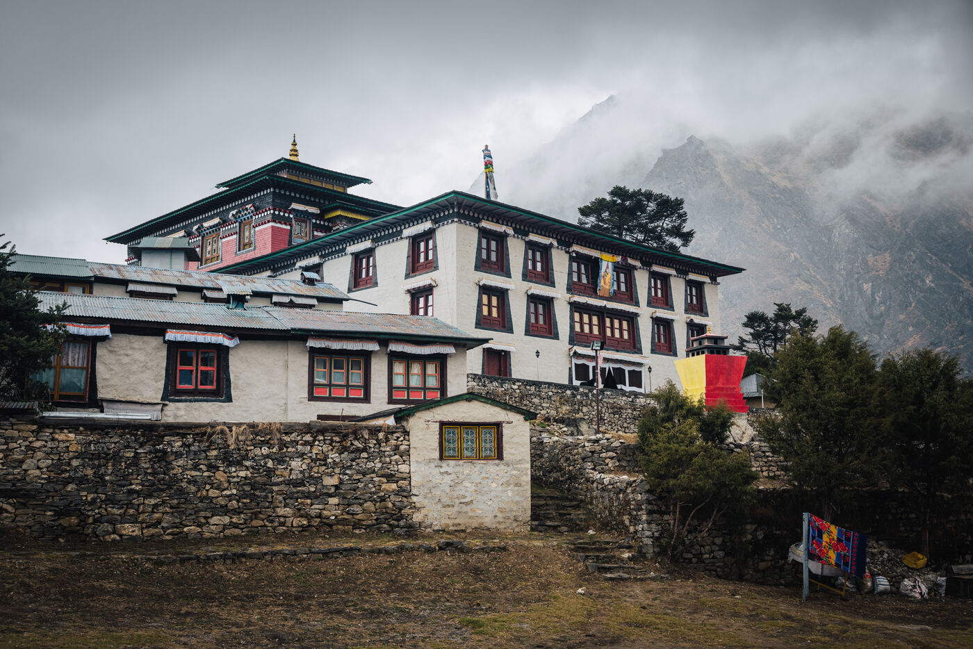 Pangboche Monastery building with ornate prayer hall roof, yellow and red banners, and mountain ridge shrouded in clouds behind