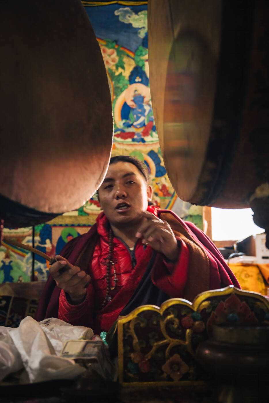 Monk in red robes holding a drumstick mid-ritual between two large ceremonial drums with a painted thangka behind
