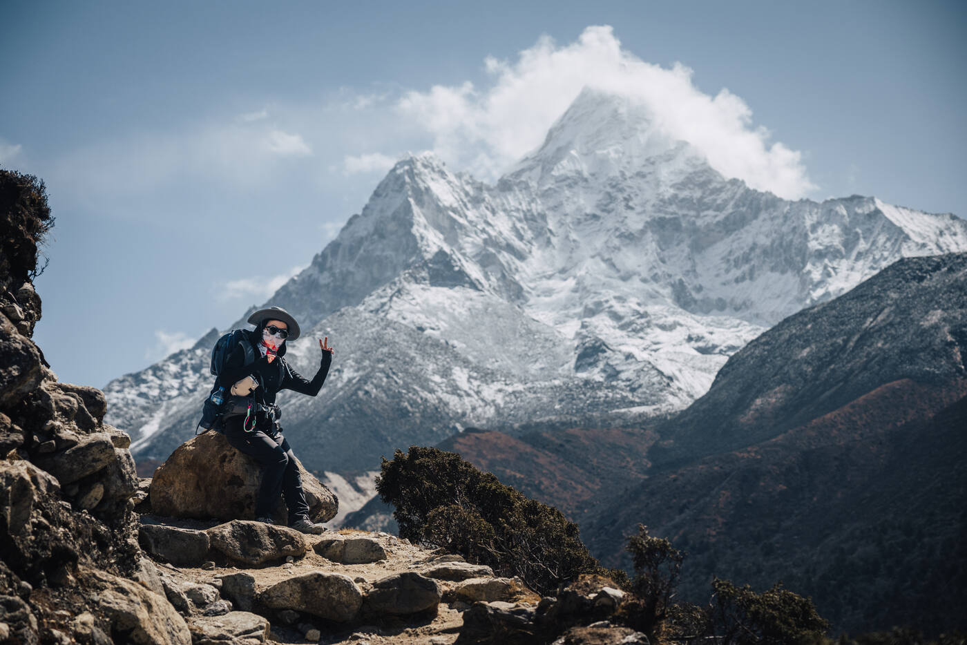 Trekker in a wide-brim hat sitting on rocks giving a peace sign with Ama Dablam rising behind