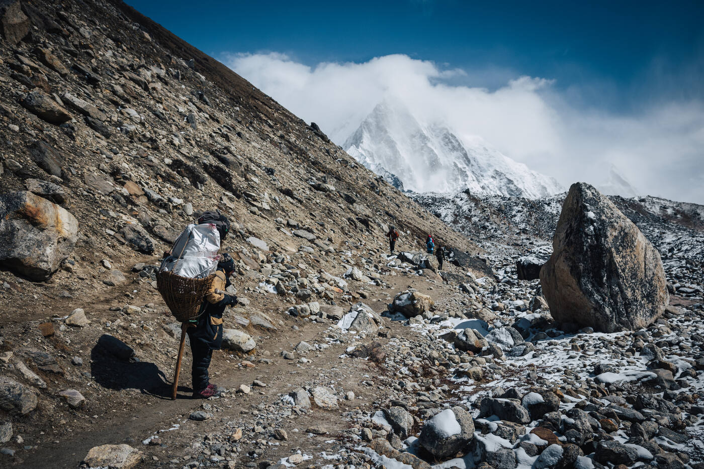 A porter with a tall woven basket crossing the high rocky moraine, a snow peak emerging from the clouds beyond