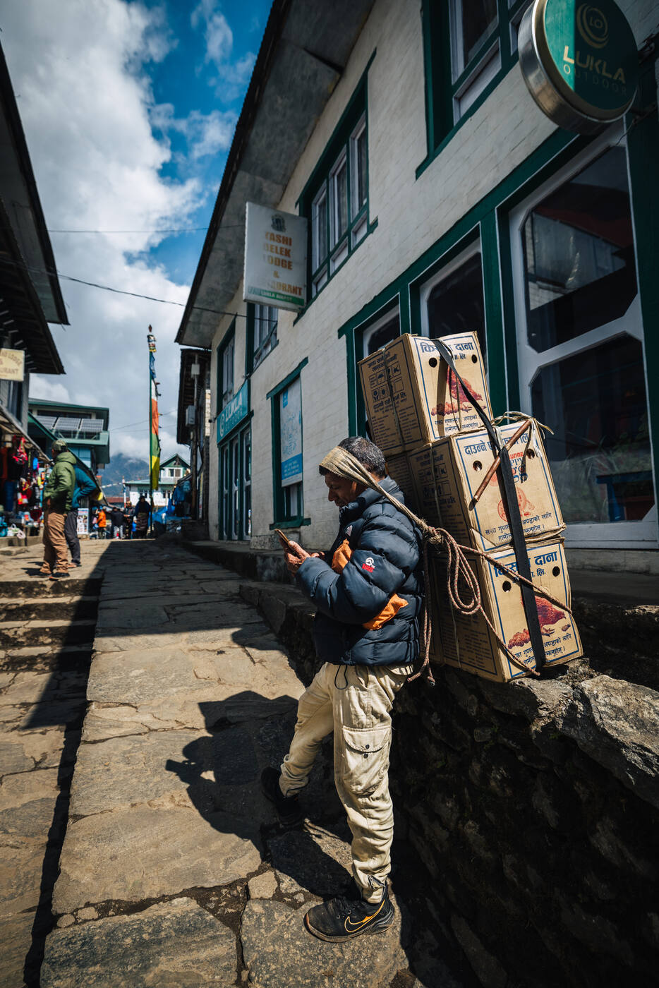 A porter in Lukla leaning against a lodge wall with stacked cardboard boxes of supplies strapped to his back