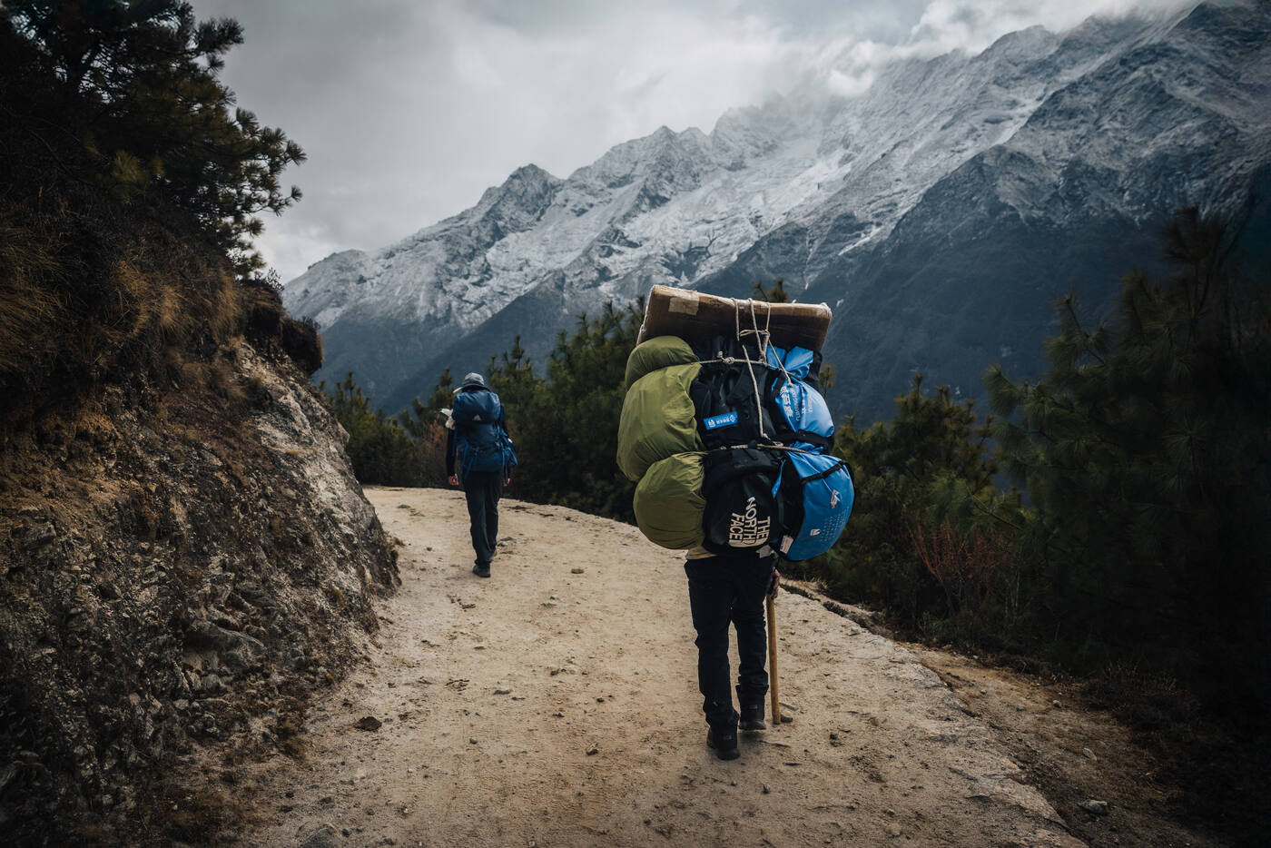 A porter with a massive bundled load walking the wide Tengboche trail toward snow peaks ahead
