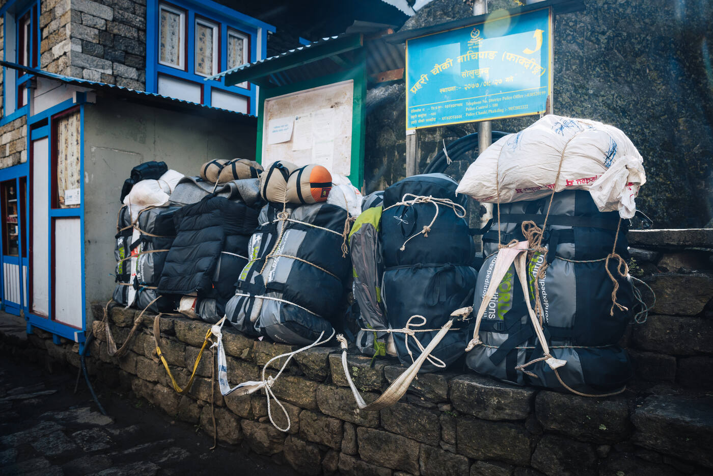 A row of porter loads — duffles wrapped in tarps and tied with rope — stacked against a stone wall outside a checkpoint