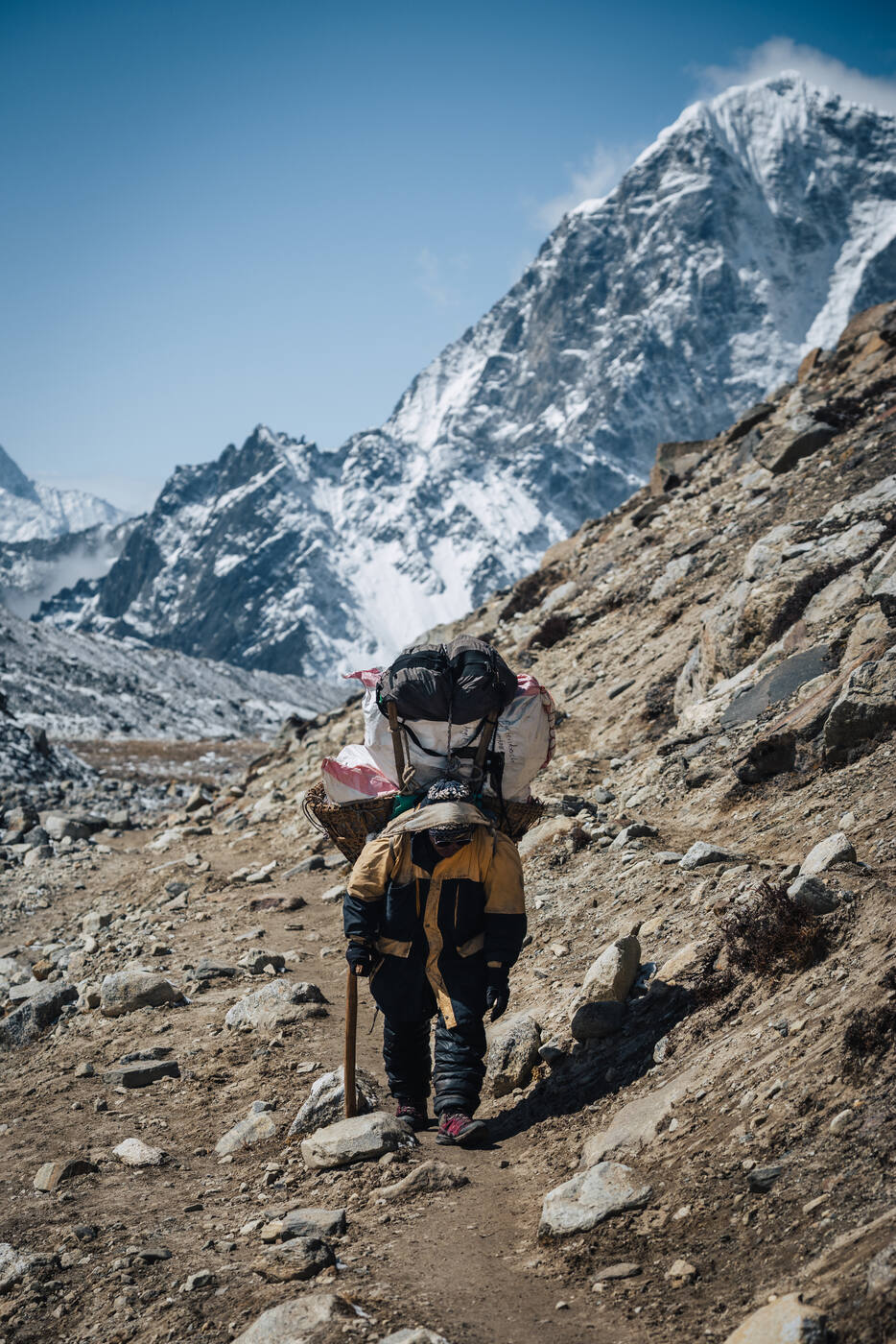 A heavily-loaded porter walking the rocky trail toward Gorak Shep, snow peaks rising sharply behind