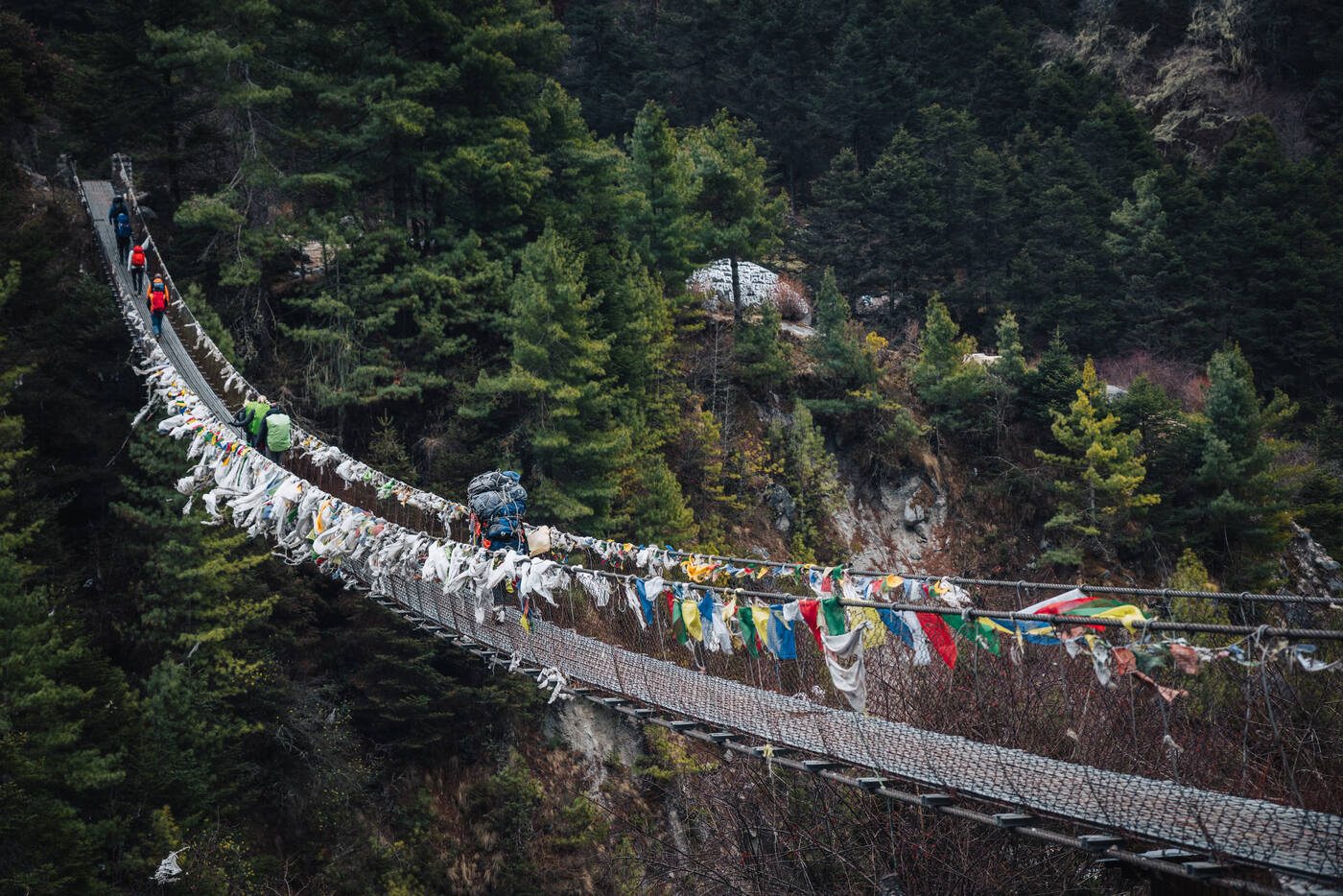 Porters crossing a long suspension bridge draped in colorful prayer flags through pine forest