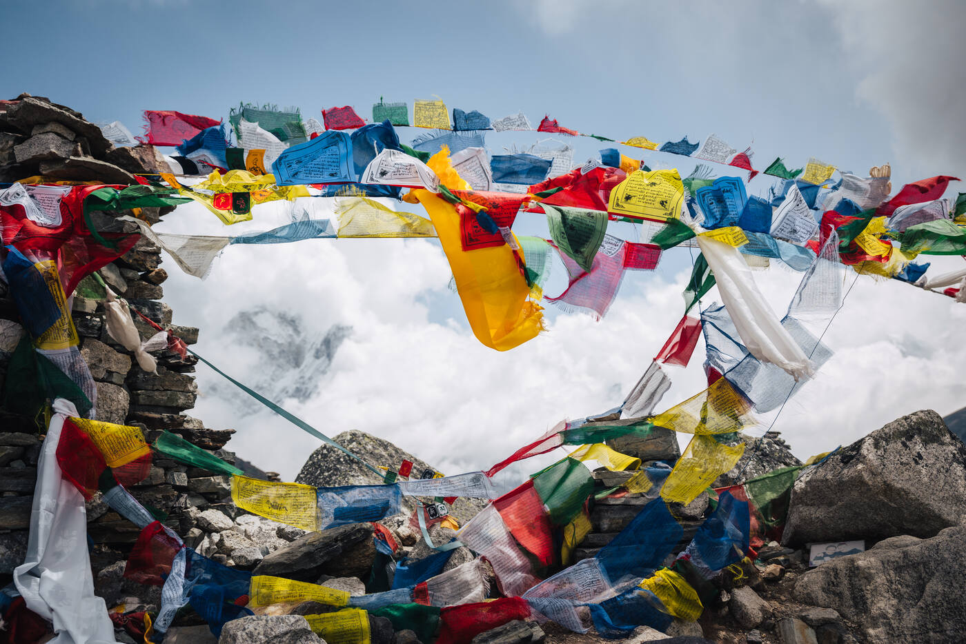 Layers of colorful prayer flags strung over the stone memorial cairns at Thukla Pass, cloud-filled sky behind