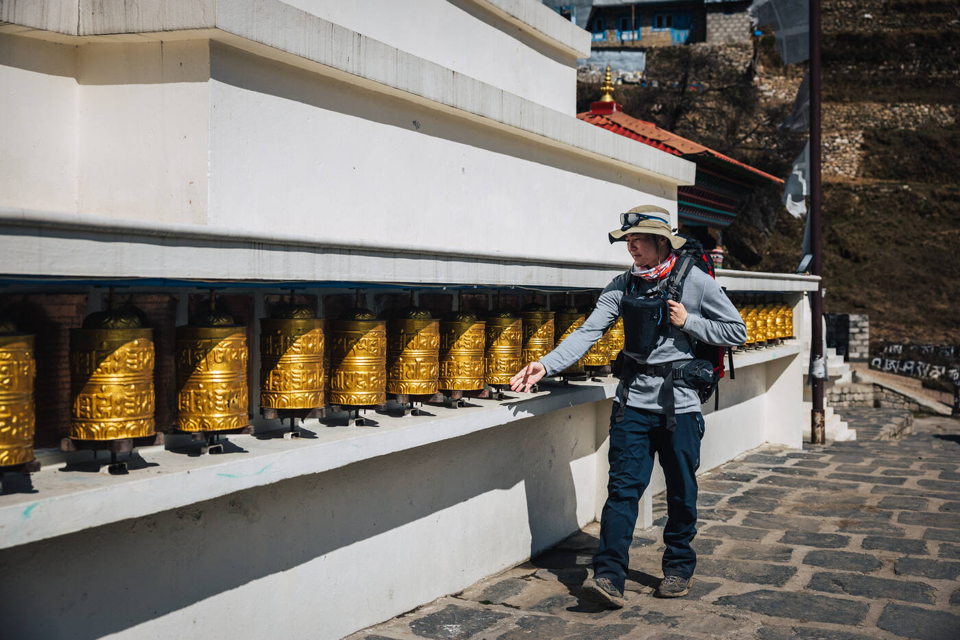 A trekker in a gray jacket and wide-brim hat spinning a long row of golden prayer wheels beside a white stupa