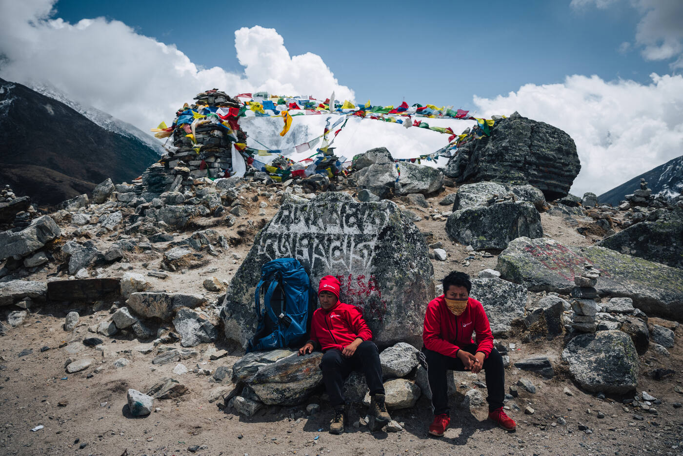 Two Sherpa porters in red jackets resting against an inscribed memorial stone at Chukpi Lhara, prayer flags strung above