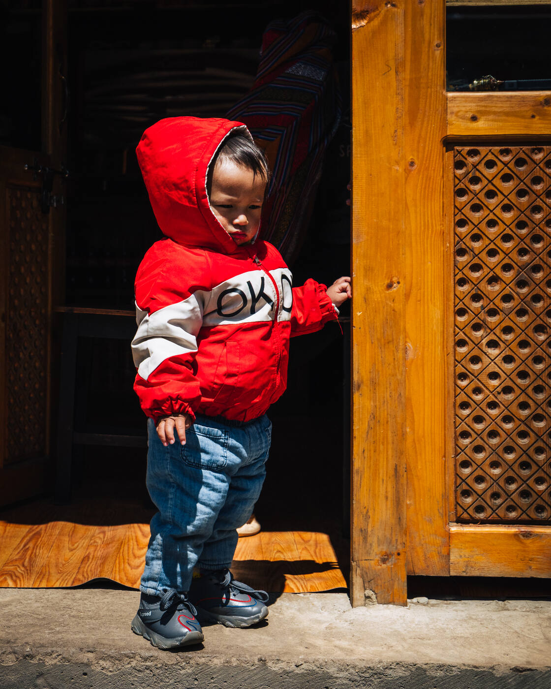 A young Sherpa child in a red hooded puffer jacket standing in a lodge doorway in Namche