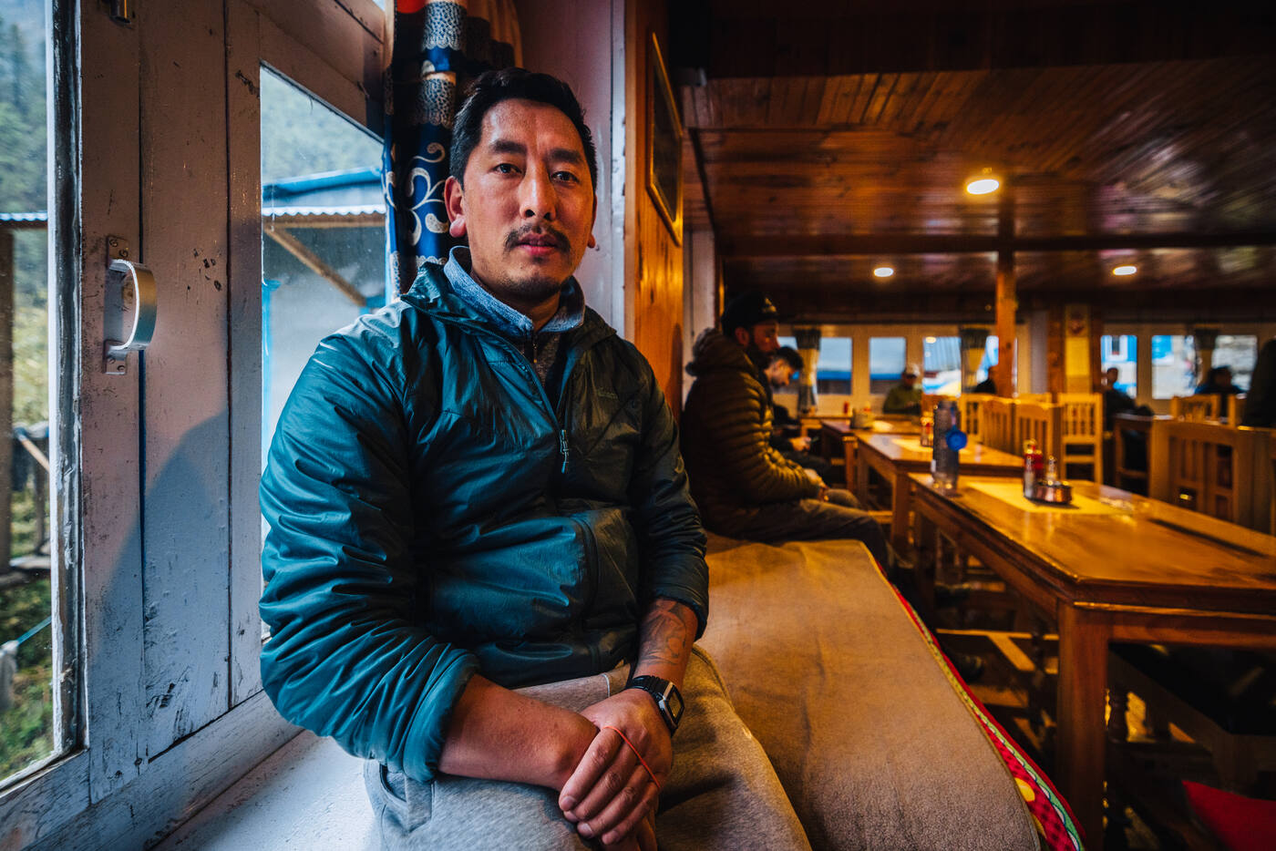 Portrait of a Sherpa guide in a teal puffy jacket inside a wood-paneled lodge dining room