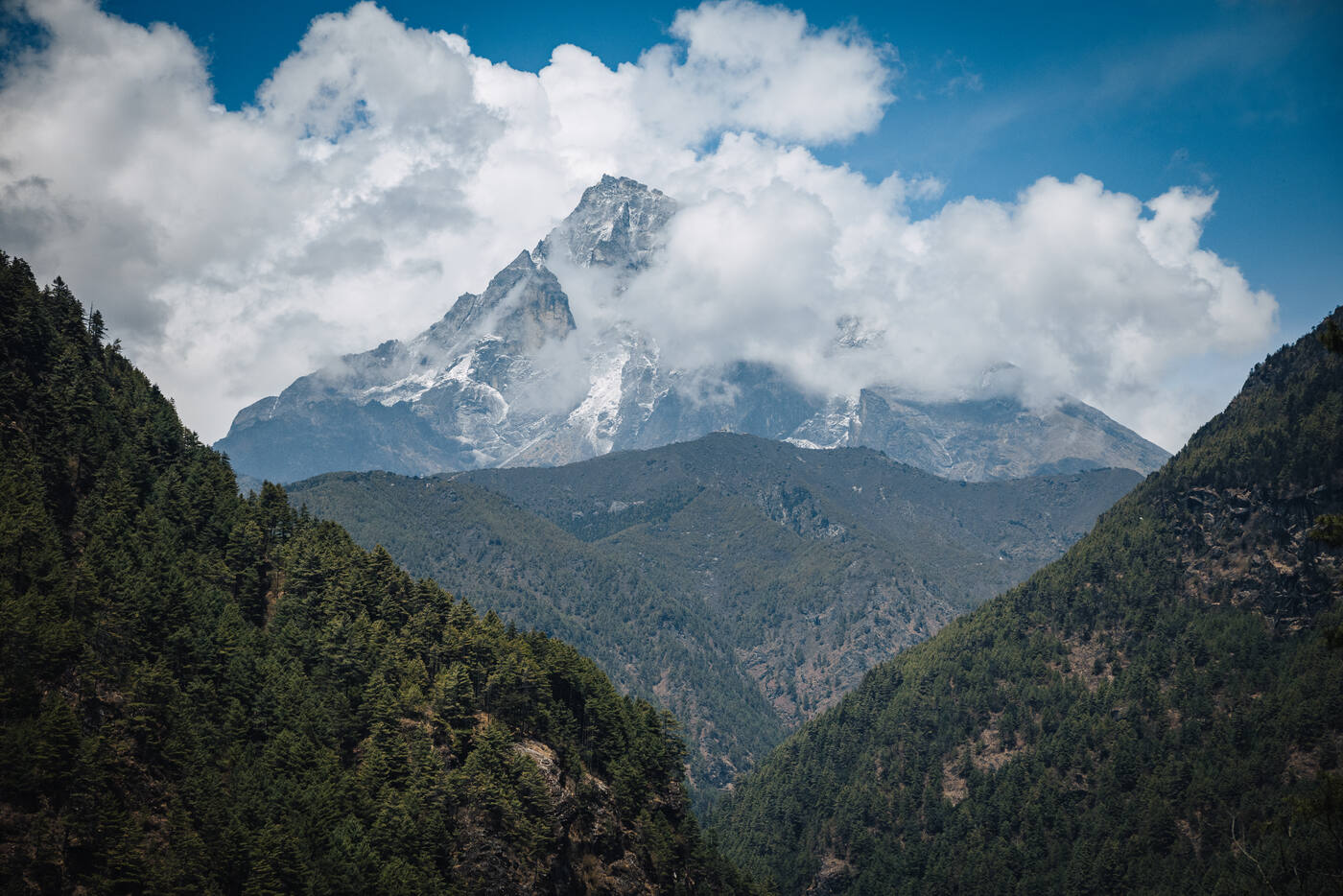 A snow-capped peak rising above forested ridges with clouds wrapping the summit, viewed from the Phakding–Namche trail