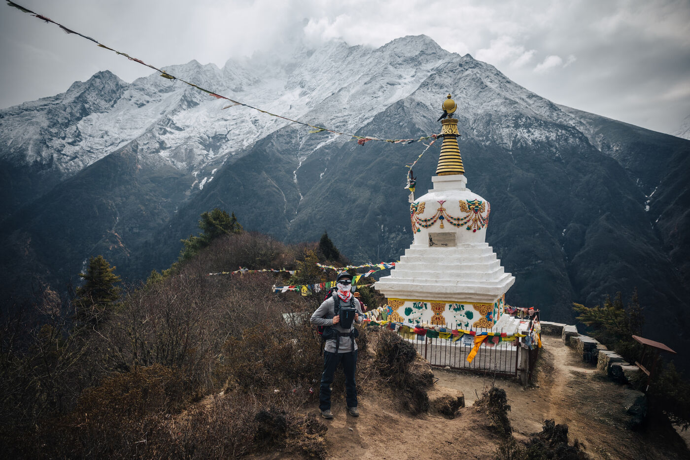 A trekker in a gray jacket and wide-brim hat standing beside a tall white-and-gold stupa on a ridge, strings of prayer flags and a massive snow-covered mountain wall rising behind
