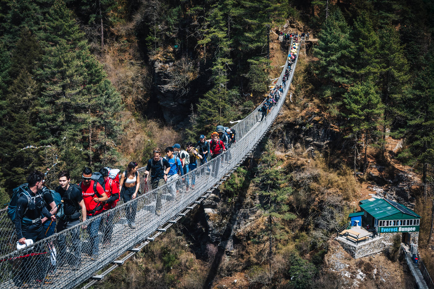 A long line of trekkers crossing a high suspension bridge above the Everest Bungee station, deep forested gorge below