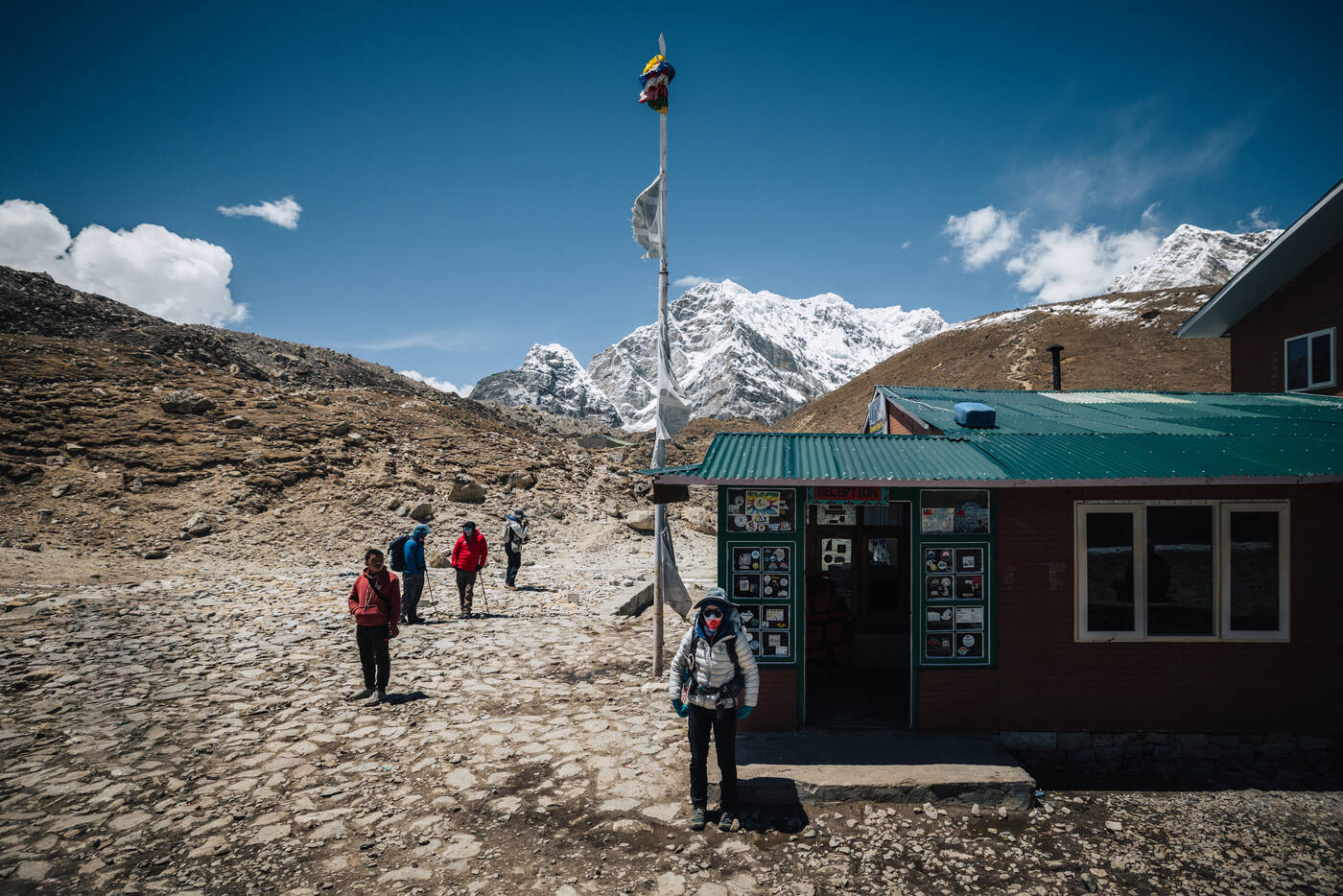 A red-roofed teahouse at Lobuche with prayer flags on a pole, snow peaks rising close behind
