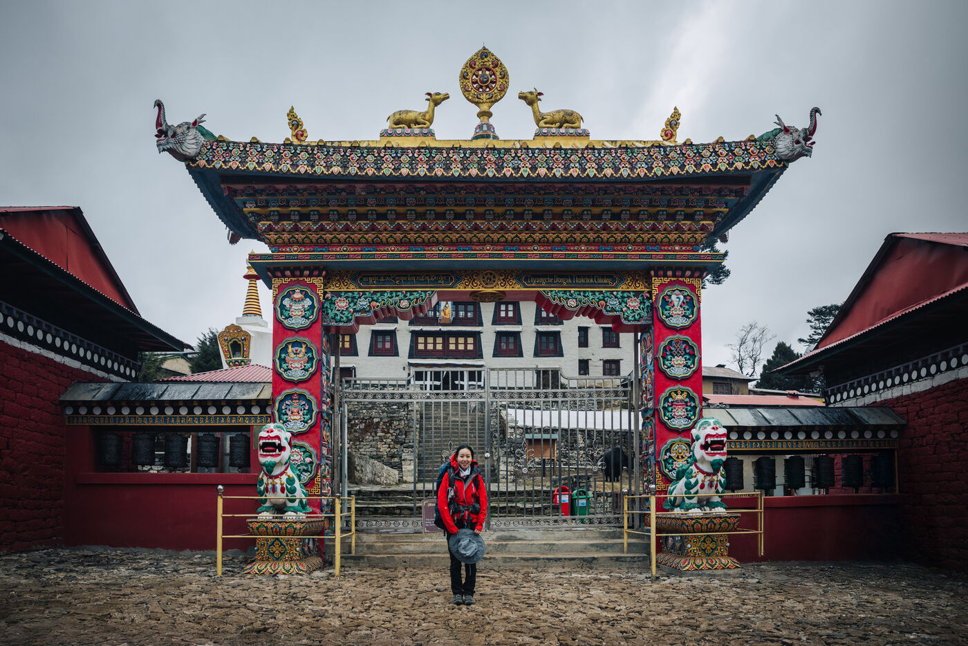 The ornate Tengboche Monastery gate with painted snow lions on either side, Grace in a red jacket standing in the courtyard