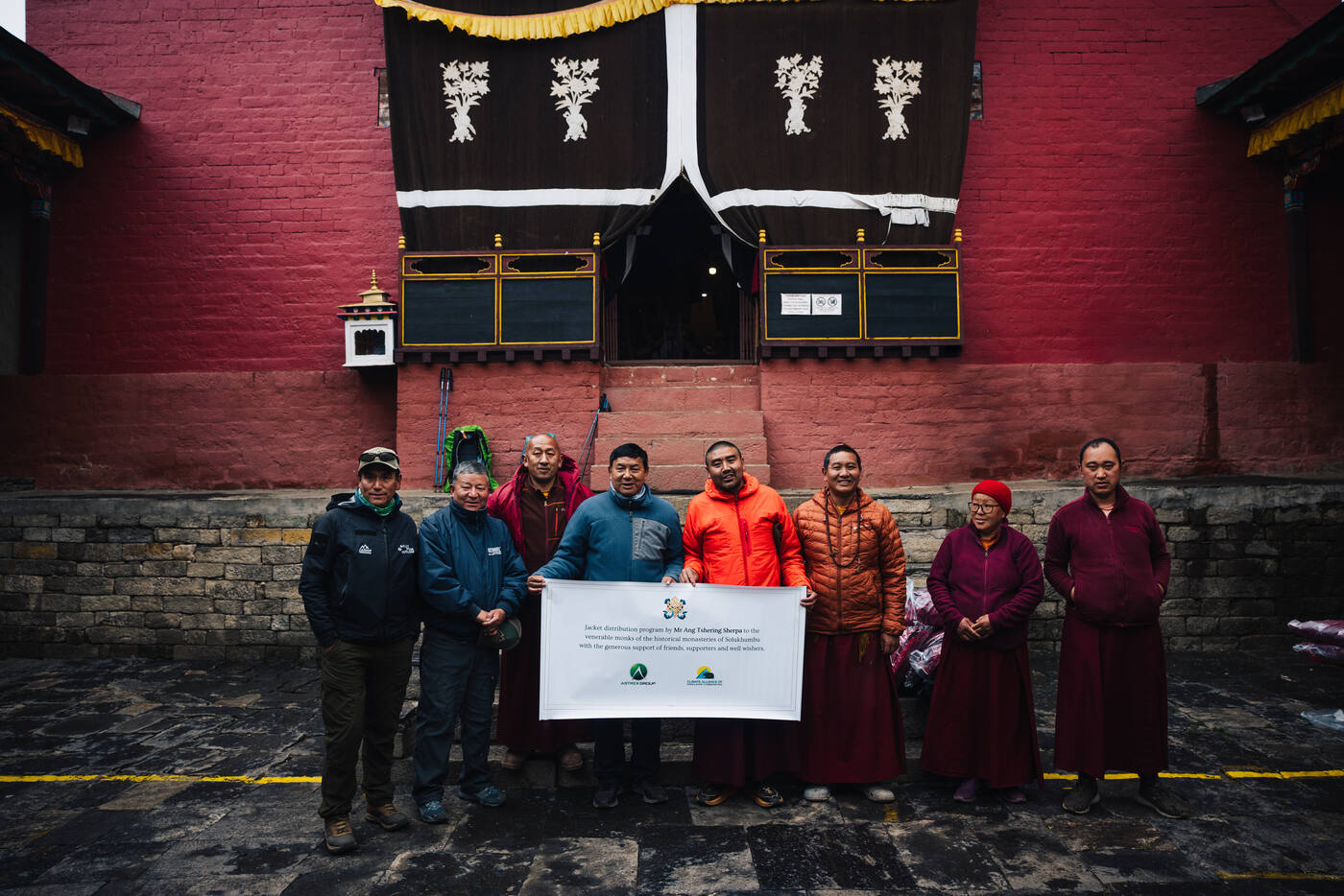 Group photo outside Tengboche Monastery — two trekkers and seven Nepali monks and staff holding a banner