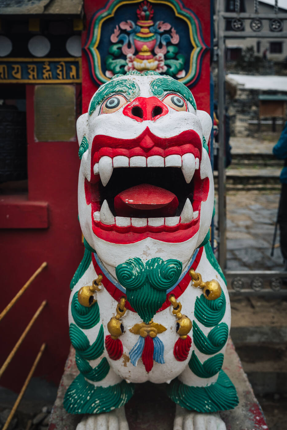 Close-up of a painted snow-lion guardian statue flanking the Tengboche monastery gate — red mouth, green mane, gold bells