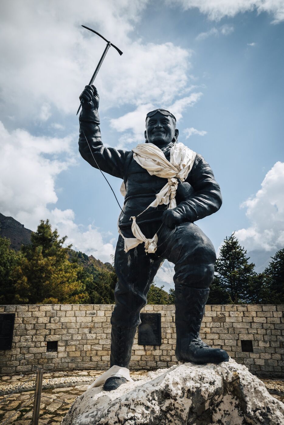 Close-up of the bronze Tenzing Norgay statue from below, ice axe raised against a cloudy sky above Namche Bazaar