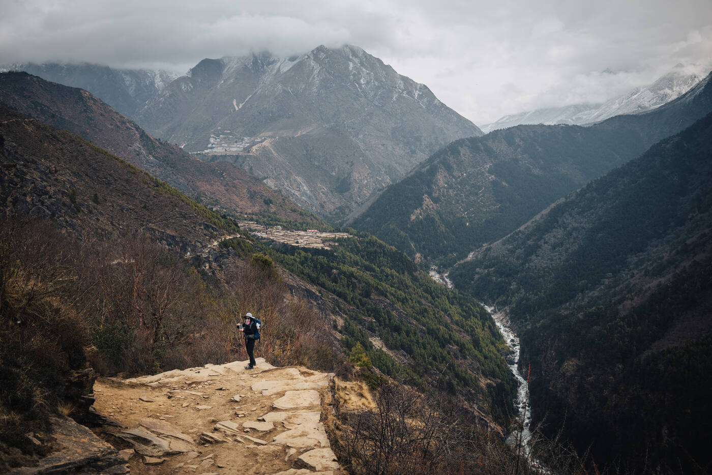 Solo trekker pausing on a rocky promontory high above the Dudh Koshi valley, river snaking through the gorge toward cloud-wrapped peaks in the distance