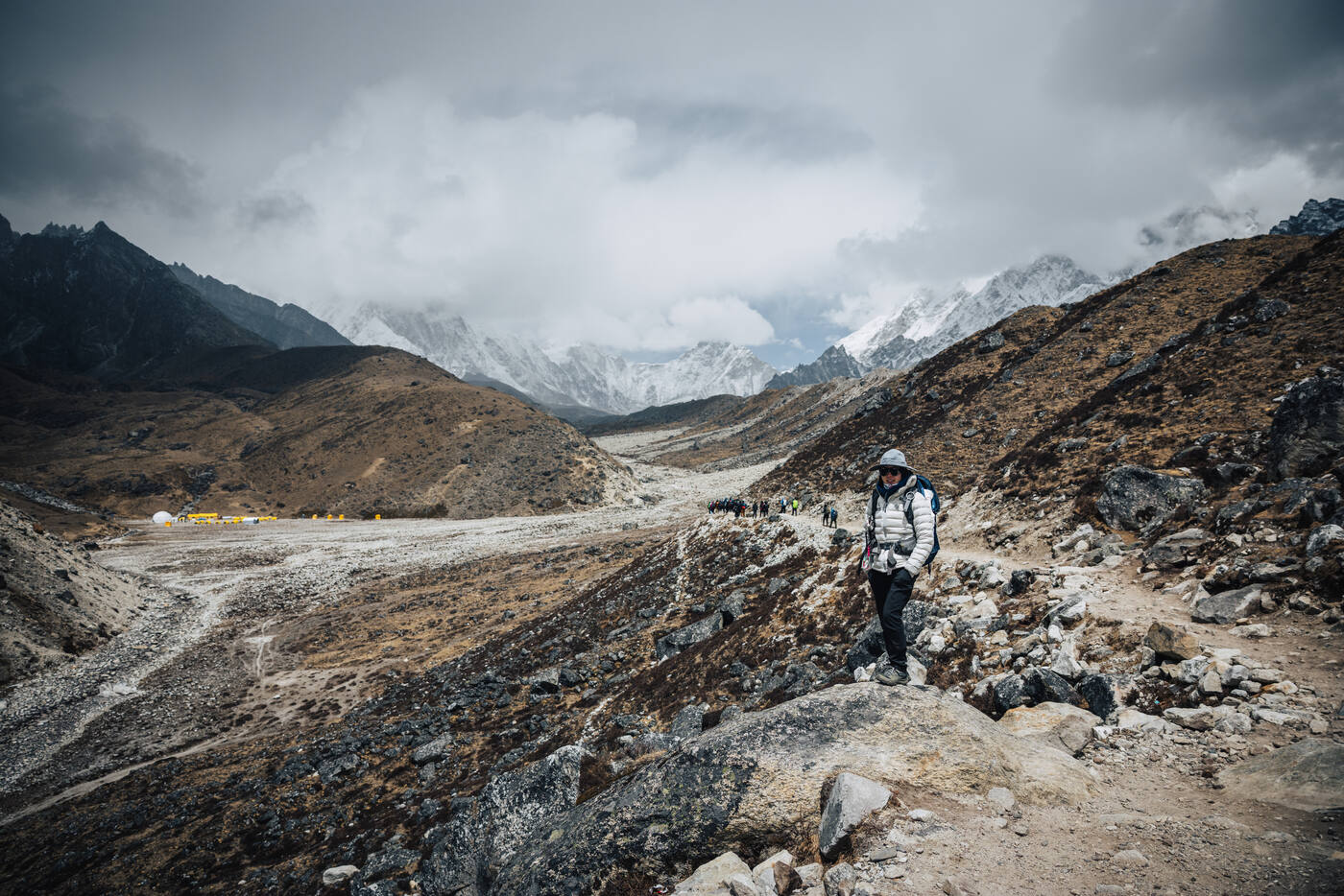 Trekker in a white puffy descending a rocky trail toward a cluster of yellow-roofed lodges on the valley floor, cloud-wrapped peaks on the horizon