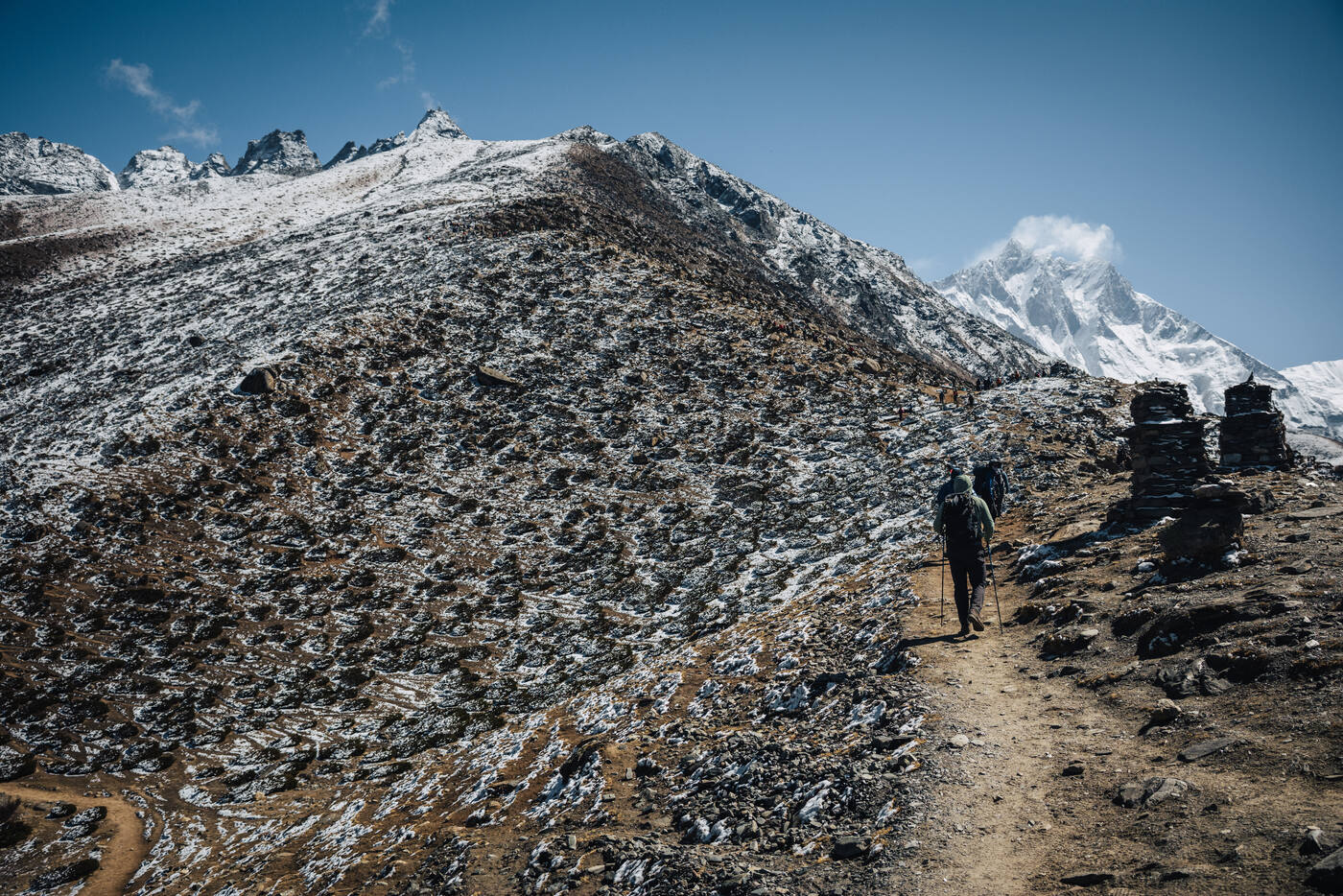 Trekker alone on a snow-dusted moraine trail with stone cairns on the right, sharp peaks behind