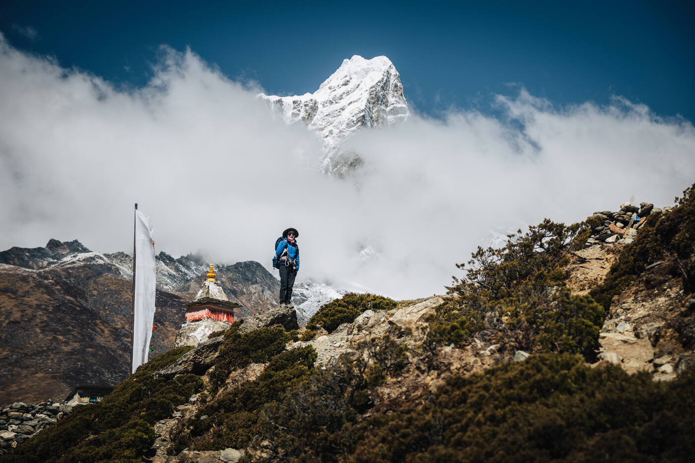 A trekker silhouetted on a rocky ridge above the chorten, snow peak emerging from thick cloud