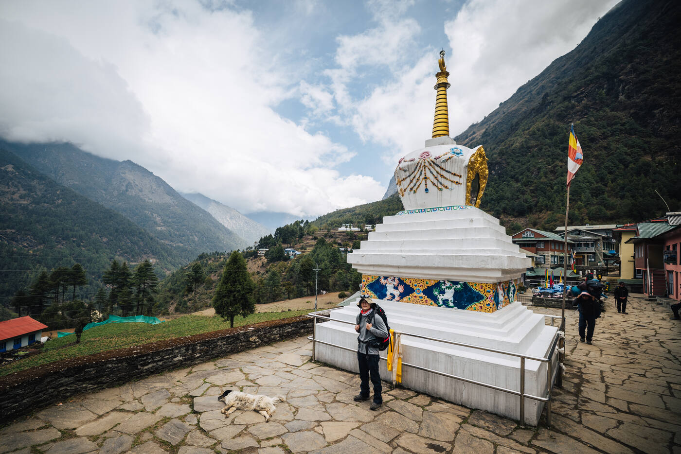 A trekker beside a tall white-and-gold stupa in a stone-paved village square, dog sleeping at the base