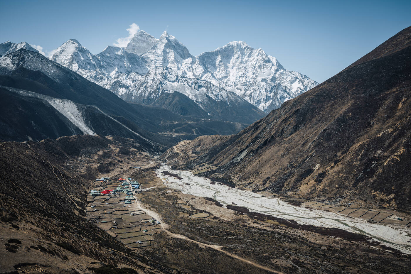 Looking down from the trail on a village beside a wide braided riverbed, terraced fields and snow peaks rising behind