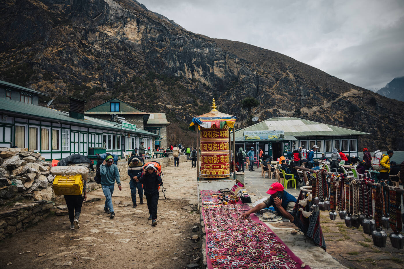 A high-altitude village square with a colorful prayer wheel at the center, vendor stalls of textiles and yak bells lining the path