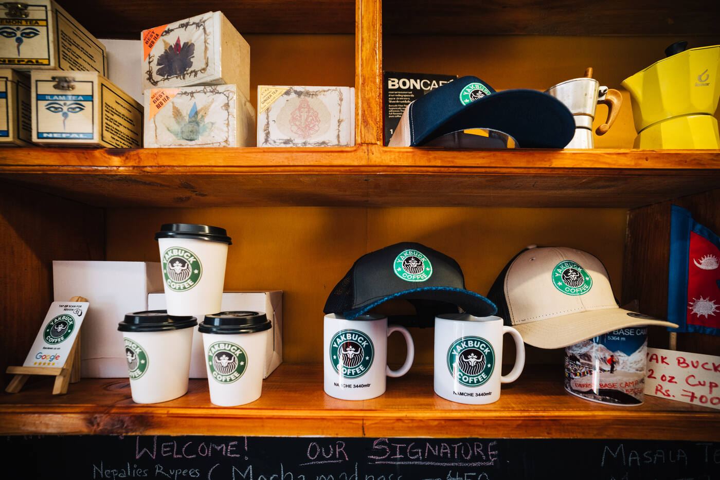 The Yakbuck Coffee merchandise shelf — branded paper cups, ceramic mugs, and ball caps lined up against a wooden cabinet