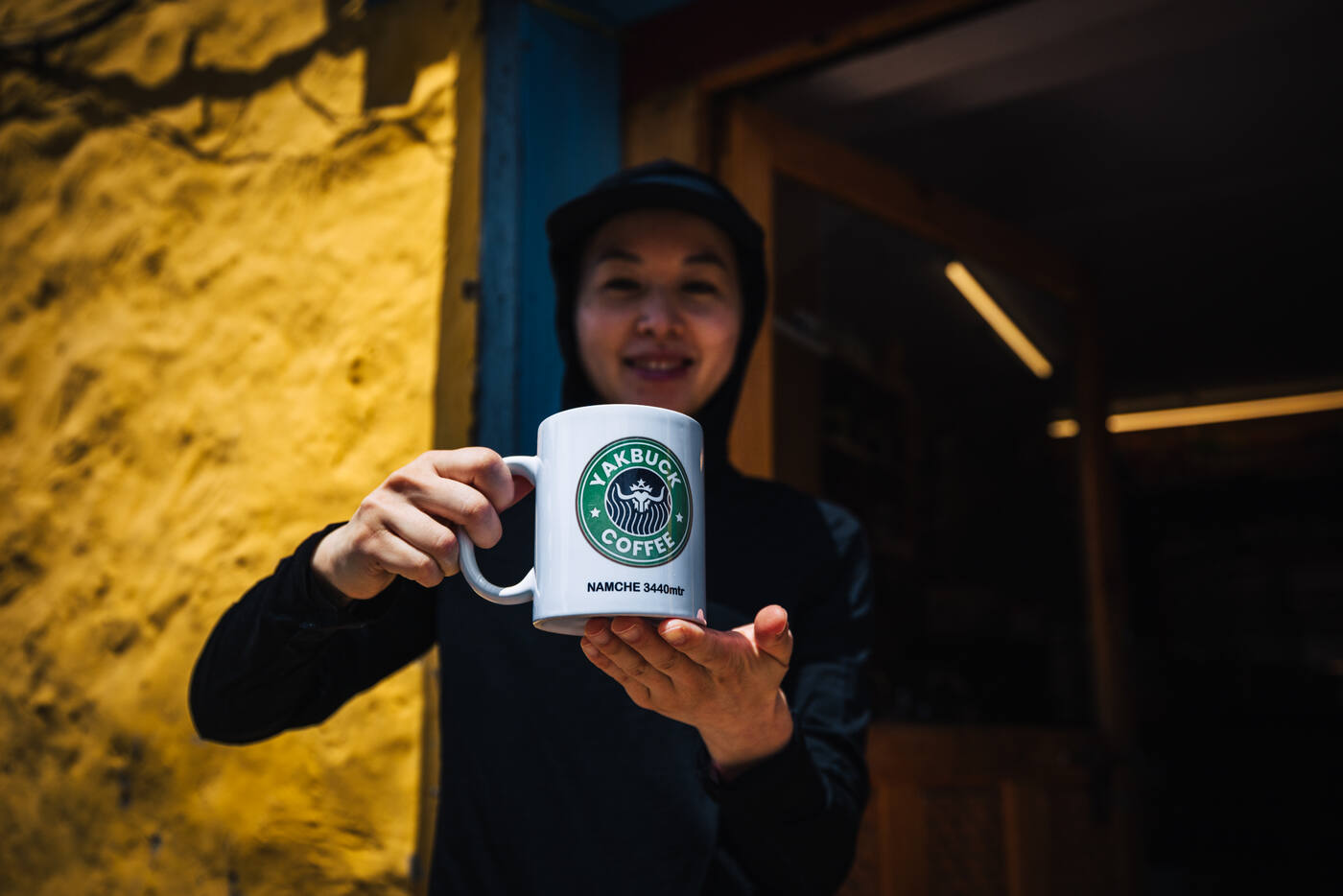 A smiling trekker holding up a Yakbuck Coffee mug printed NAMCHE 3440mtr against a yellow lodge wall