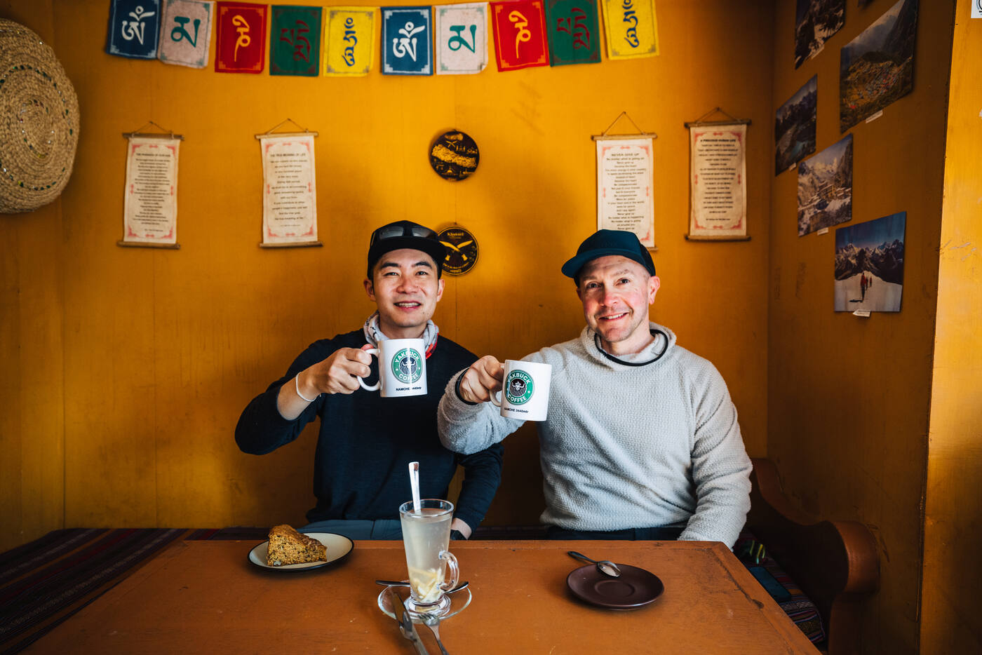 Jason and a friend at a Yakbuck Coffee table clinking branded mugs over a lemon-ginger drink, prayer flags strung above