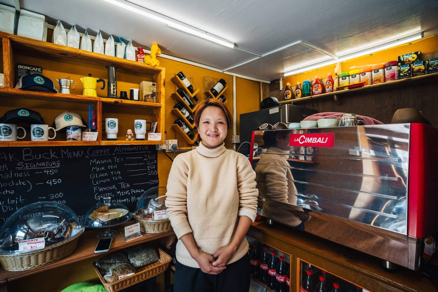 The Yakbuck Coffee owner smiling behind her counter — menu chalkboard, La Cimbali espresso machine, branded mugs on shelves